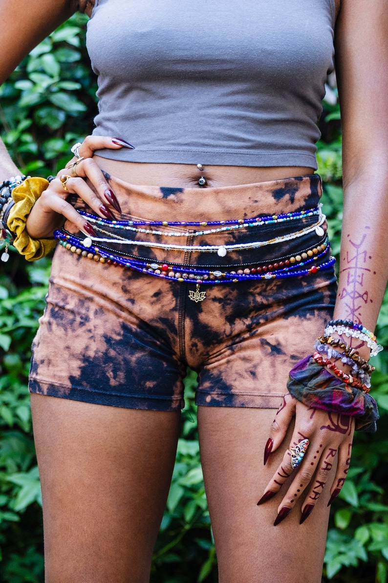 Puede incluir: Una persona lleva un top gris sin mangas y pantalones cortos de te&ntilde;ido anudado con m&uacute;ltiples cadenas de cintura de cuentas. La persona tiene tatuajes de henna en los brazos y las manos, y lleva varias pulseras en una mu&ntilde;eca.