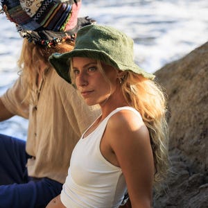 May include: A woman wearing a green bucket hat and a white tank top. The hat has a wide brim and a textured appearance. The woman has long blonde hair and is looking off to the side. Another person is visible in the background wearing a patterned head covering.