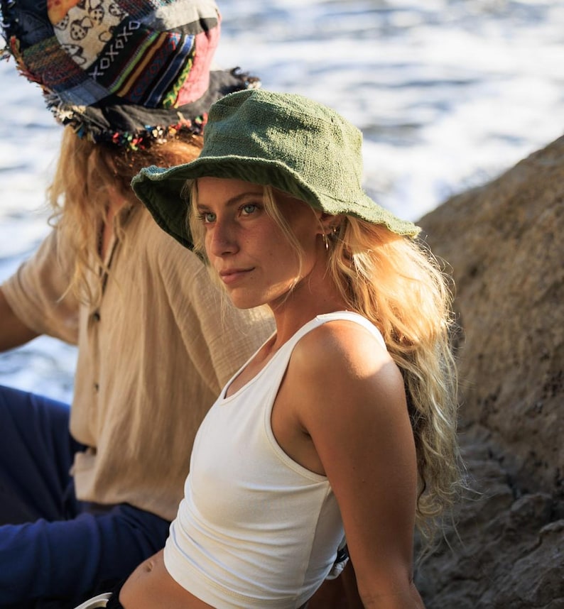 May include: A woman wearing a green bucket hat and a white tank top. The hat has a wide brim and a textured appearance. The woman has long blonde hair and is looking off to the side. Another person is visible in the background wearing a patterned head covering.