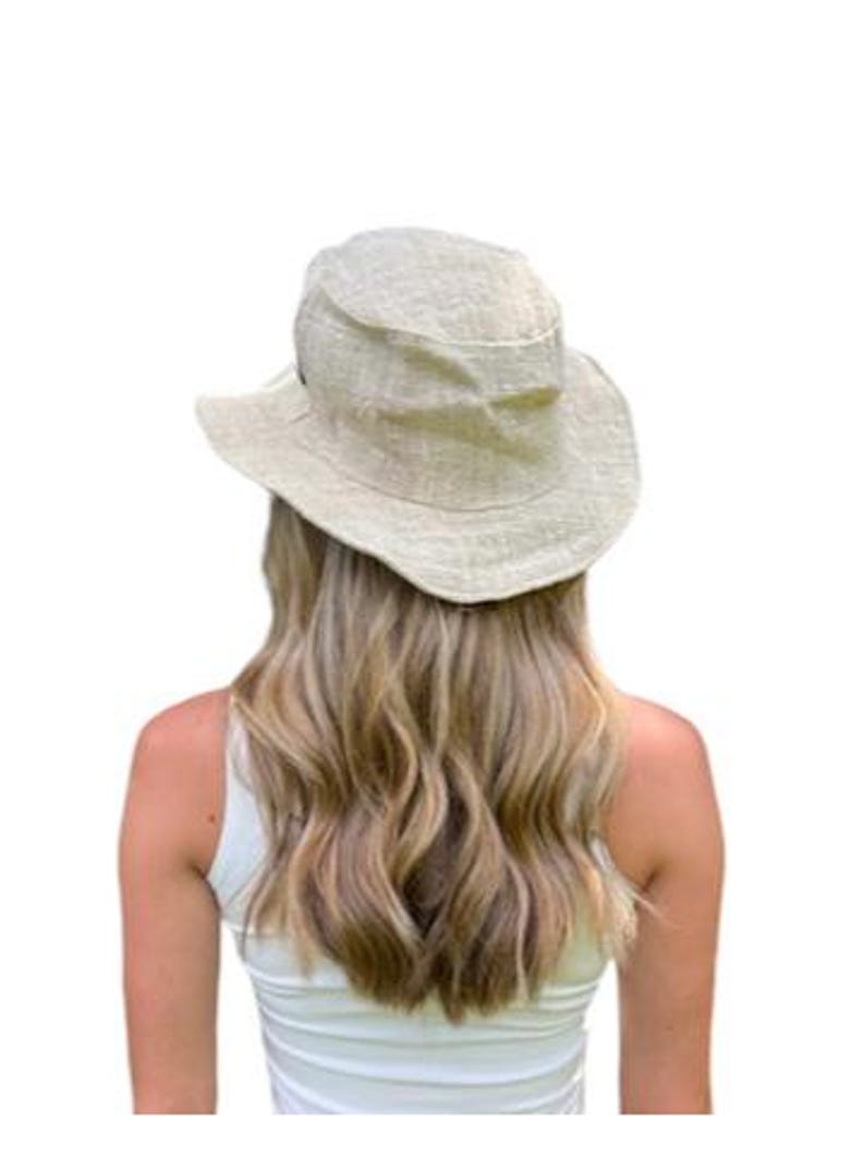 May include: A light beige sun hat with a wide brim. The hat is made of a textured fabric and has a slightly rounded crown. The hat is worn by a person wearing a white tank top.