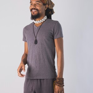 May include: A dark gray V-neck t-shirt and matching dark gray drawstring pants. The outfit is accessorized with a brown patterned headscarf, a white cowrie shell choker, a long dark pendant necklace, and a multi-layered brown wooden bead bracelet.