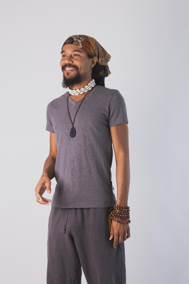 May include: A dark gray V-neck t-shirt and matching dark gray drawstring pants. The outfit is accessorized with a brown patterned headscarf, a white cowrie shell choker, a long dark pendant necklace, and a multi-layered brown wooden bead bracelet.