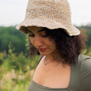 May include: A textured bucket hat crafted from a blend of tan and white fibers, featuring a rounded crown and a short, slightly flared brim. The hat displays a visible woven construction with a variegated color pattern throughout the material.
