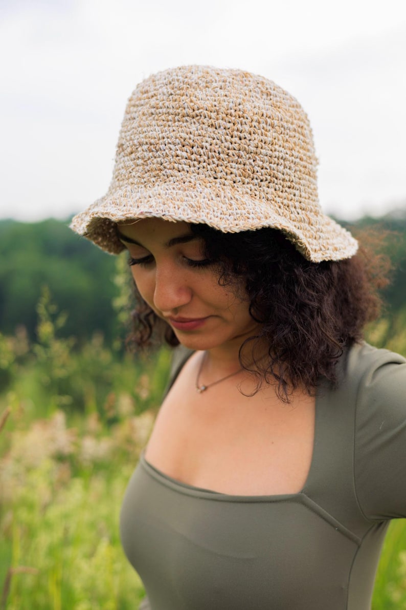 May include: A textured bucket hat crafted from a blend of tan and white fibers, featuring a rounded crown and a short, slightly flared brim. The hat displays a visible woven construction with a variegated color pattern throughout the material.