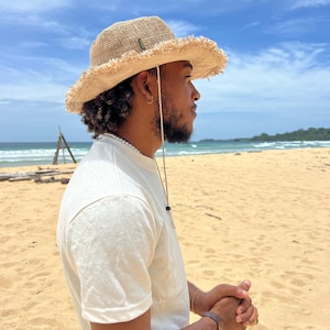 May include: A person wearing a light brown, wide-brimmed straw hat with a frayed edge, a white t-shirt, and a brown bracelet on their wrist. They are standing on a sandy beach with a blue sky and ocean in the background.