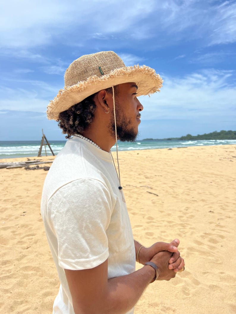 May include: A person wearing a light brown, wide-brimmed straw hat with a frayed edge, a white t-shirt, and a brown bracelet on their wrist. They are standing on a sandy beach with a blue sky and ocean in the background.