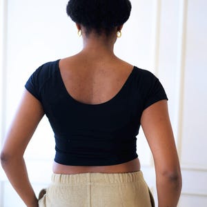 Op de afbeelding: Een zwarte crop top met een ronde hals en korte mouwen. Het model draagt een beige joggingbroek. De persoon heeft haar haar in een knot en draagt gouden oorringen.