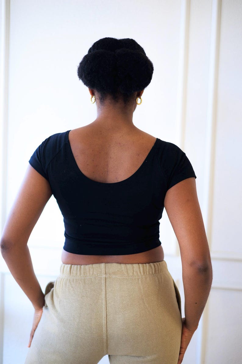 Op de afbeelding: Een zwarte crop top met een ronde hals en korte mouwen. Het model draagt een beige joggingbroek. De persoon heeft haar haar in een knot en draagt gouden oorringen.