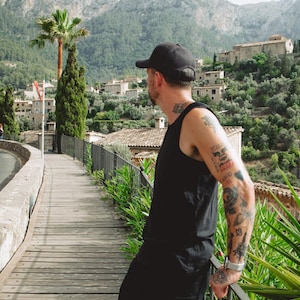 Puede incluir: Una gorra de b&eacute;isbol negra, una camiseta sin mangas negra y pantalones cortos negros usados por una persona con tatuajes en el brazo y el cuello, de pie en una pasarela de madera con vistas a un pueblo de monta&ntilde;a con edificios de piedra.