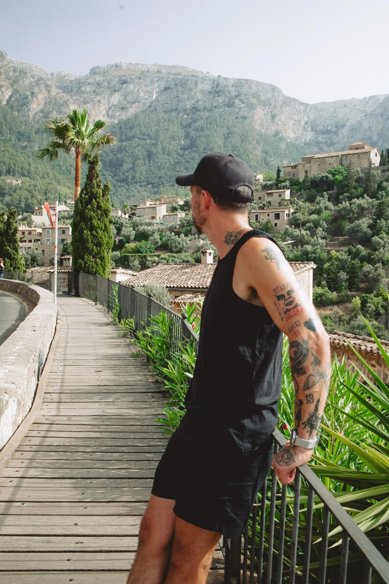 Puede incluir: Una gorra de b&eacute;isbol negra, una camiseta sin mangas negra y pantalones cortos negros usados por una persona con tatuajes en el brazo y el cuello, de pie en una pasarela de madera con vistas a un pueblo de monta&ntilde;a con edificios de piedra.