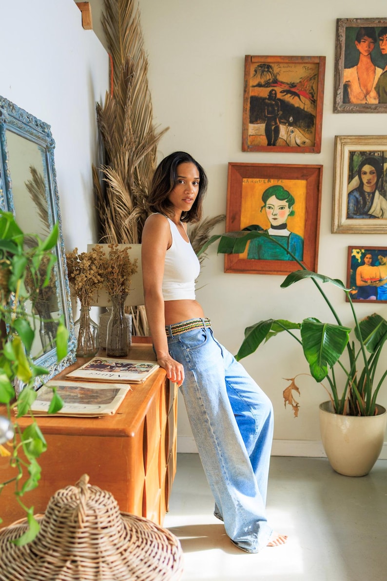May include: A white sleeveless crop top paired with light blue denim jeans featuring a woven multicolored belt. The jeans have a wide-leg silhouette and a classic five-pocket design, sitting low on the waist with a visible button and zipper fly.