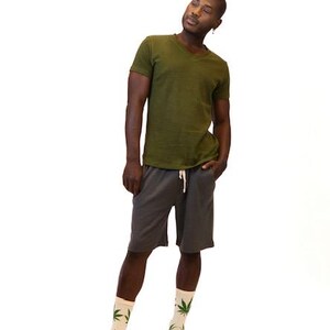 May include: An olive green V-neck t-shirt, charcoal gray shorts with a white drawstring, and cream-colored socks featuring a repeating green leaf pattern. The clothing items are worn together as a complete outfit against a plain white background.