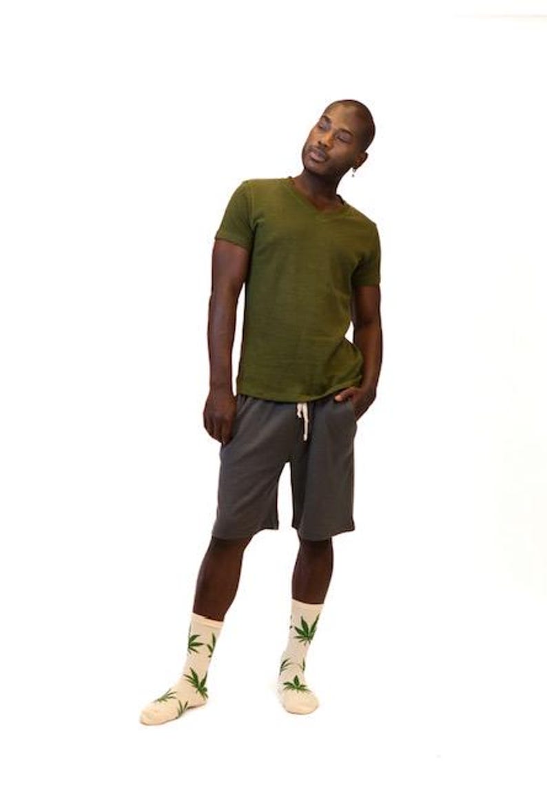 May include: An olive green V-neck t-shirt, charcoal gray shorts with a white drawstring, and cream-colored socks featuring a repeating green leaf pattern. The clothing items are worn together as a complete outfit against a plain white background.
