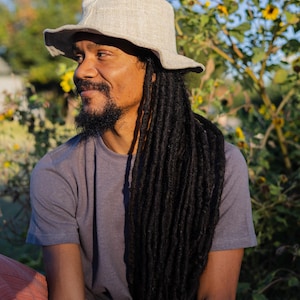 May include: A light gray, wide-brimmed hat with a slightly textured appearance. The hat is worn by a person with long, dark dreadlocks and a gray t-shirt. The background features yellow flowers and greenery under a blue sky.