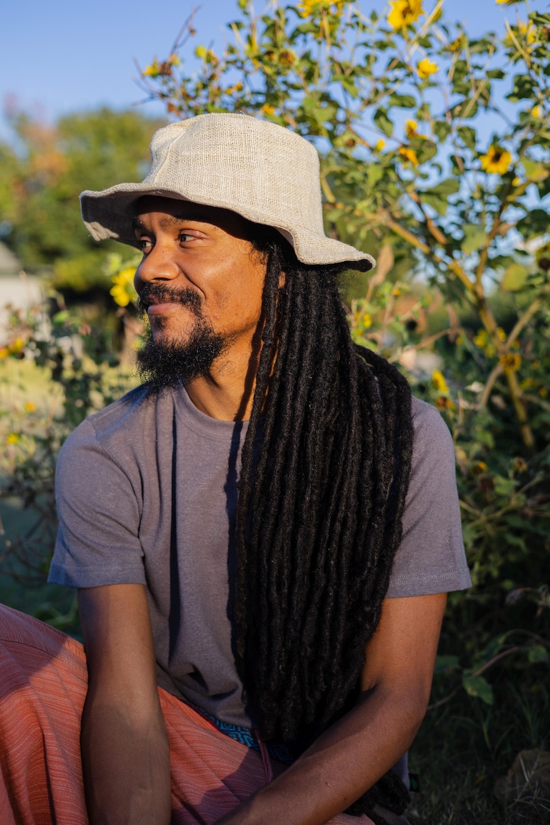 May include: A light gray, wide-brimmed hat with a slightly textured appearance. The hat is worn by a person with long, dark dreadlocks and a gray t-shirt. The background features yellow flowers and greenery under a blue sky.