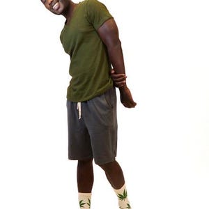 May include: An olive green V-neck t-shirt, dark gray shorts with a white drawstring, and beige crew socks featuring a repeating green cannabis leaf pattern. The clothing items are worn together as a complete outfit against a plain white background.