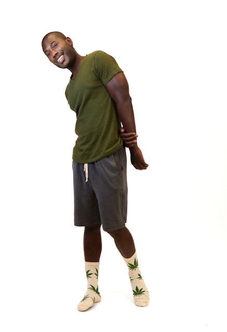 May include: An olive green V-neck t-shirt, dark gray shorts with a white drawstring, and beige crew socks featuring a repeating green cannabis leaf pattern. The clothing items are worn together as a complete outfit against a plain white background.
