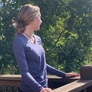 May include: A woman wearing a long-sleeved, dark blue v-neck shirt stands on a wooden deck, looking out at a green tree line.