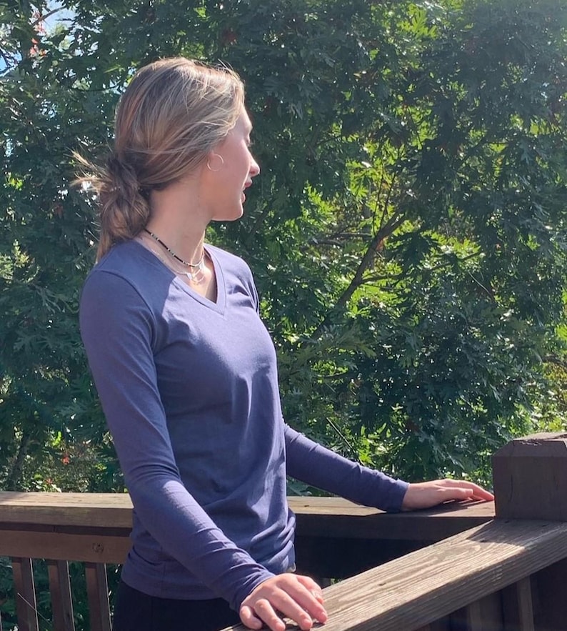 May include: A woman wearing a long-sleeved, dark blue v-neck shirt stands on a wooden deck, looking out at a green tree line.