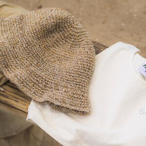 May include: A beige crochet bucket hat, a white sleeveless top with a tag reading "Asaee M", and a tan fabric piece. The white top features the text "SAVING THE PLANET one leaf at a time" in white and green lettering on the lower right side.