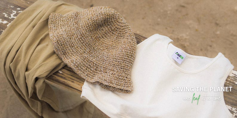 May include: A beige crochet bucket hat, a white sleeveless top with a tag reading "Asaee M", and a tan fabric piece. The white top features the text "SAVING THE PLANET one leaf at a time" in white and green lettering on the lower right side.