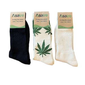 May include: Three pairs of crew socks with cardboard labels reading Asatre Saving the planet, one leaf at a time! One pair is solid black, one pair is off-white with a green cannabis leaf pattern, and one pair is solid off-white. All socks are ribbed.