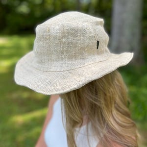 May include: A beige, wide-brimmed sun hat made of woven material. The hat has a slightly crumpled appearance and a small black tag on the side. The hat is designed to provide shade from the sun.