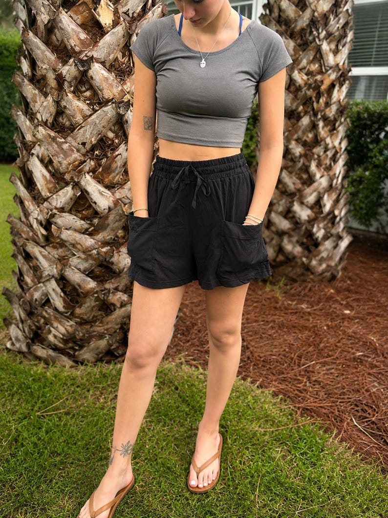 May include: A charcoal gray short-sleeved crop top with a scoop neckline paired with black high-waisted shorts featuring an elastic drawstring waistband and two large front patch pockets. The shorts have a relaxed fit and a mid-thigh length.