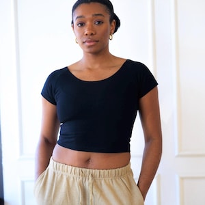 Op de afbeelding: Een zwarte crop top met korte mouwen en een beige joggingbroek met zakken. De top heeft een ronde hals. De joggingbroek heeft een taille met trekkoord. De persoon draagt gouden oorringen.