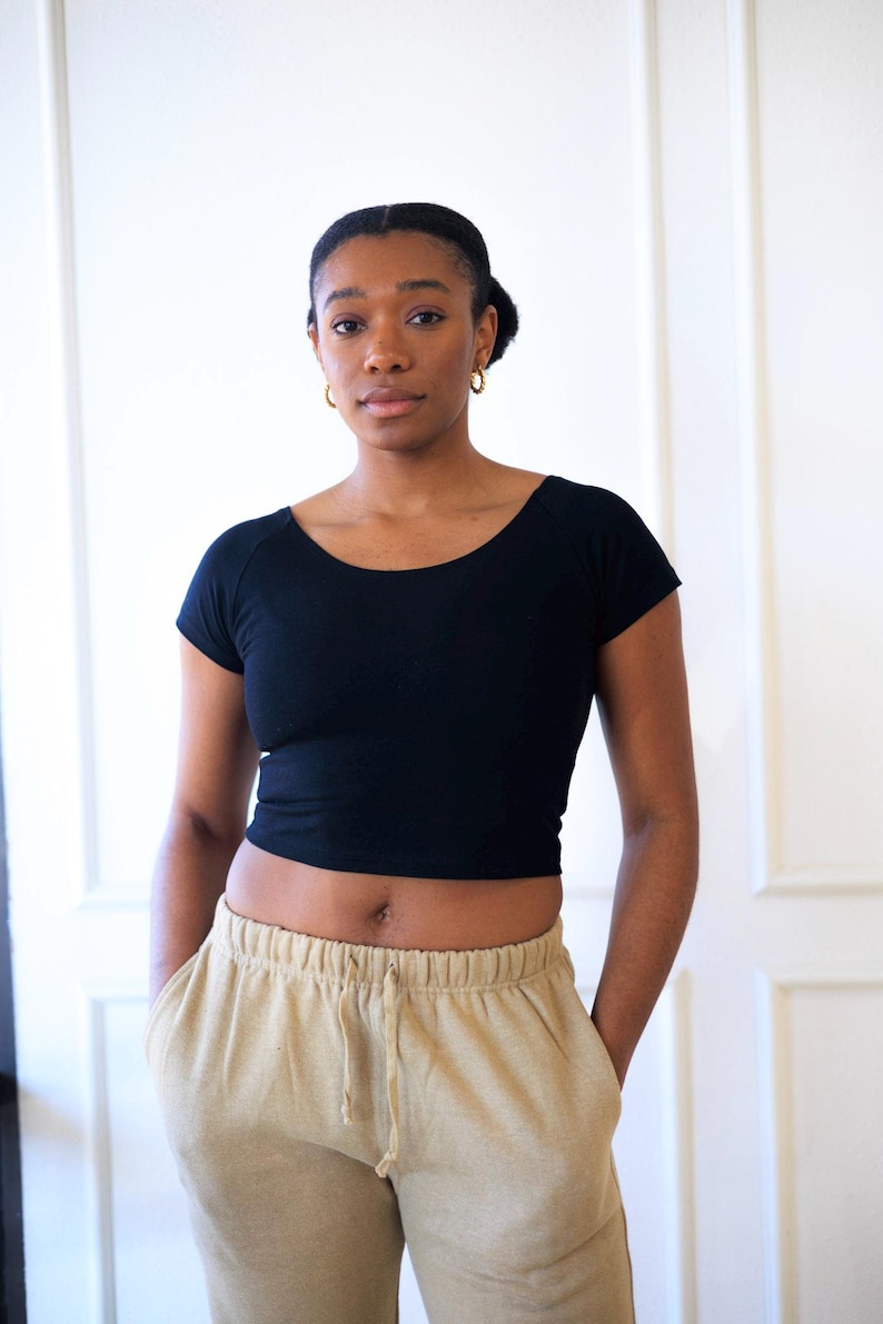 Op de afbeelding: Een zwarte crop top met korte mouwen en een beige joggingbroek met zakken. De top heeft een ronde hals. De joggingbroek heeft een taille met trekkoord. De persoon draagt gouden oorringen.