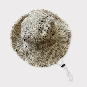 May include: A wide-brimmed, natural-colored hemp hat with a frayed edge and a drawstring. The hat is made of a woven material and has a simple design.