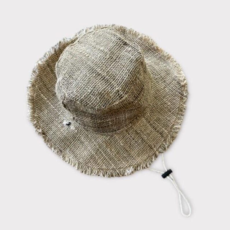 May include: A wide-brimmed, natural-colored hemp hat with a frayed edge and a drawstring. The hat is made of a woven material and has a simple design.