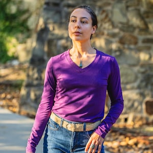 May include: A woman wearing a purple long-sleeved shirt with a v-neck and a brown belt. She is wearing blue jeans and has a necklace with a green stone pendant.