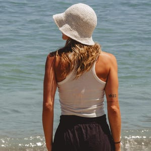 May include: A cream-colored crochet bucket hat worn with a white ribbed tank top and a black skirt. The hat features a textured, woven pattern with a downward-sloping brim, while the tank top has a racerback design and a fitted silhouette.