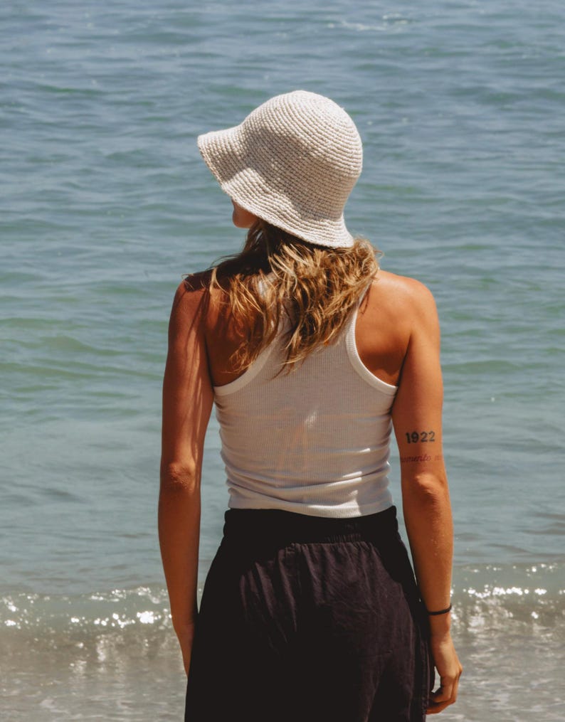 May include: A cream-colored crochet bucket hat worn with a white ribbed tank top and a black skirt. The hat features a textured, woven pattern with a downward-sloping brim, while the tank top has a racerback design and a fitted silhouette.