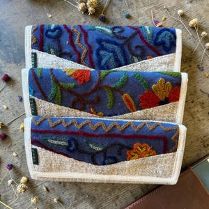 Könnte beinhalten: Drei blaue, bestickte Clutch-Geldbörsen mit floralen und geometrischen Designs. Die Geldbörsen haben eine beige Tasche und weiße Zierleisten. Das Wort "HEMP" ist auf der Seite jeder Geldbörse zu sehen. Die Geldbörsen sind auf einer Holzoberfläche gestapelt.