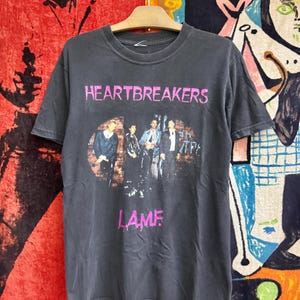 Puede incluir: Una camiseta de manga corta de color gris carbón con un estampado gráfico de una banda. El texto HEARTBREAKERS está impreso en rosa sobre los miembros de la banda, y la palabra L.A.M.F. está impresa en rosa debajo de la imagen en la parte frontal.