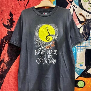 May include: Black t-shirt featuring a graphic from "The Nightmare Before Christmas." The design includes a yellow moon, a silhouette of Jack Skellington, and the film's title in white lettering. The shirt is hanging on a wooden hanger.