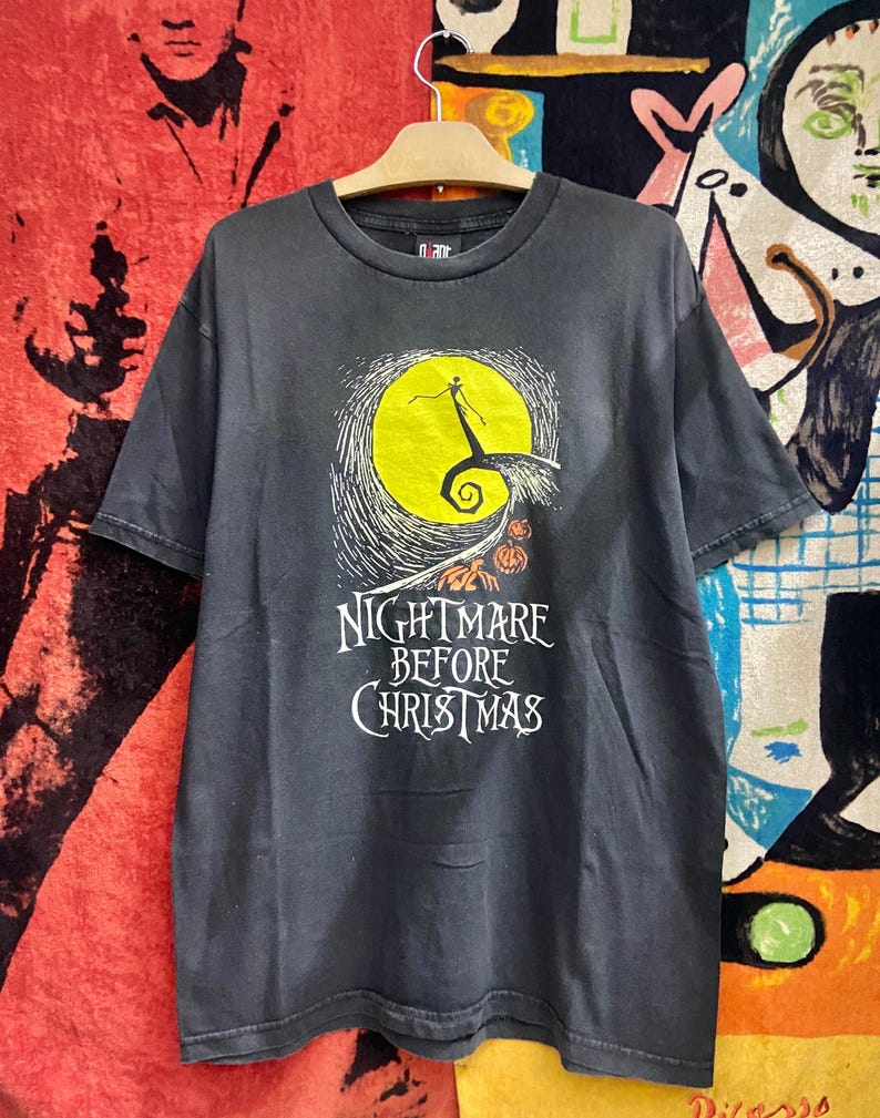 May include: Black t-shirt featuring a graphic from "The Nightmare Before Christmas." The design includes a yellow moon, a silhouette of Jack Skellington, and the film's title in white lettering. The shirt is hanging on a wooden hanger.