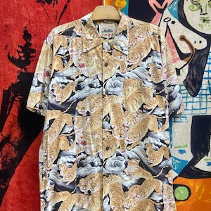 Puede incluir: Camisa de manga corta con botones y un estampado repetido de tigres en tonos beige, marrón y blanco sobre un fondo de nubes grises. La camisa está colgada de una percha de madera.