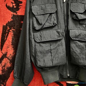 Peut inclure: Veste de bombardier gris fonc&eacute; avec plusieurs poches avant et un poignet c&ocirc;tel&eacute;. La veste a une fermeture &eacute;clair et une petite &eacute;tiquette de logo. La veste est faite d'un mat&eacute;riau l&eacute;ger, possiblement en nylon.