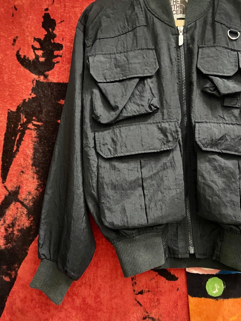 Peut inclure: Veste de bombardier gris fonc&eacute; avec plusieurs poches avant et un poignet c&ocirc;tel&eacute;. La veste a une fermeture &eacute;clair et une petite &eacute;tiquette de logo. La veste est faite d'un mat&eacute;riau l&eacute;ger, possiblement en nylon.