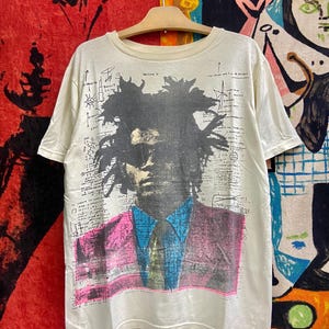 K&ouml;nnte beinhalten: Ein wei&szlig;es T-Shirt mit einem Schwarzwei&szlig;bild des K&uuml;nstlers Jean-Michel Basquiat. Das Bild ist von handgeschriebenen Notizen und Zeichnungen umgeben. Das Shirt hat einen Rundhalsausschnitt und kurze &Auml;rmel.