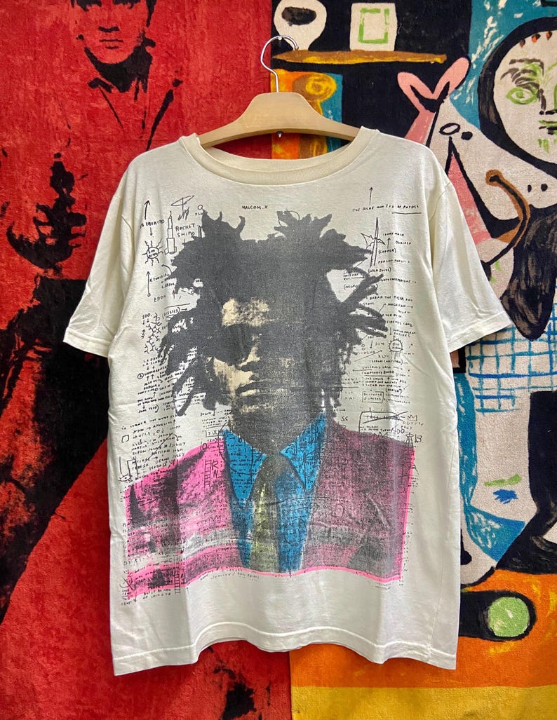 K&ouml;nnte beinhalten: Ein wei&szlig;es T-Shirt mit einem Schwarzwei&szlig;bild des K&uuml;nstlers Jean-Michel Basquiat. Das Bild ist von handgeschriebenen Notizen und Zeichnungen umgeben. Das Shirt hat einen Rundhalsausschnitt und kurze &Auml;rmel.