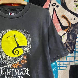 May include: Black t-shirt featuring a graphic from "The Nightmare Before Christmas." The design includes a yellow full moon, a silhouette of Jack Skellington, and the words "Nightmare Before Christmas" in white. Two orange pumpkins are also visible.