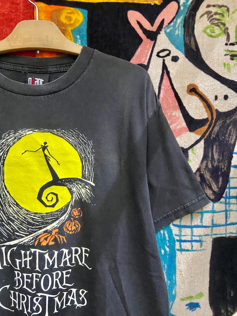 May include: Black t-shirt featuring a graphic from "The Nightmare Before Christmas." The design includes a yellow full moon, a silhouette of Jack Skellington, and the words "Nightmare Before Christmas" in white. Two orange pumpkins are also visible.