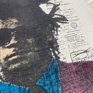 K&ouml;nnte beinhalten: Ein Vintage-T-Shirt in Wei&szlig; mit einem Schwarzwei&szlig;-Portr&auml;t von Malcolm X. Das Portr&auml;t ist von einem farbenfrohen, abstrakten Design und Text umgeben, der "Malcolm.X", "The Bilme", "Babar the Tiger", "Magellan", "Circus Came", "DUM", "Serum Cholesterol", "Vitavus Vasa", "Campiones Bonfo", "Job Louis", "Scar Pa", "(2 Asia)", "(Radium)", "5.S", "Pajaro" und "DC" beinhaltet.
