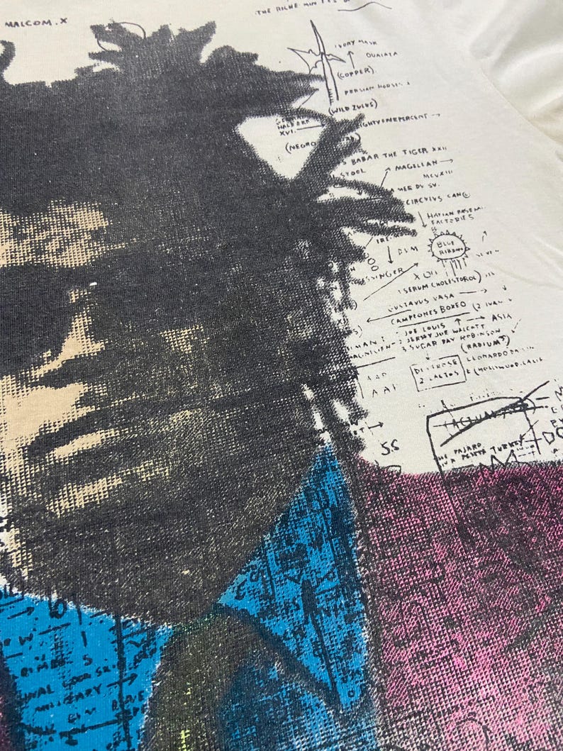 K&ouml;nnte beinhalten: Ein Vintage-T-Shirt in Wei&szlig; mit einem Schwarzwei&szlig;-Portr&auml;t von Malcolm X. Das Portr&auml;t ist von einem farbenfrohen, abstrakten Design und Text umgeben, der "Malcolm.X", "The Bilme", "Babar the Tiger", "Magellan", "Circus Came", "DUM", "Serum Cholesterol", "Vitavus Vasa", "Campiones Bonfo", "Job Louis", "Scar Pa", "(2 Asia)", "(Radium)", "5.S", "Pajaro" und "DC" beinhaltet.