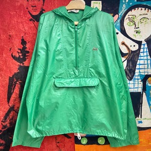 May include: A bright green hooded anorak jacket with a half-zip closure and a front pocket. The jacket features the Lacoste logo on the chest and a drawstring at the hem. The anorak is hanging on a wooden hanger.