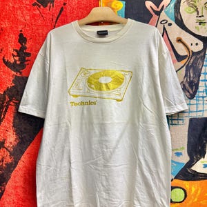 Puede incluir: Una camiseta de manga corta y cuello redondo en color blanco roto con un gráfico dorado de un tocadiscos Technics en el pecho. La palabra Technics está impresa en dorado debajo de la ilustración del tocadiscos. Etiqueta negra en el cuello.